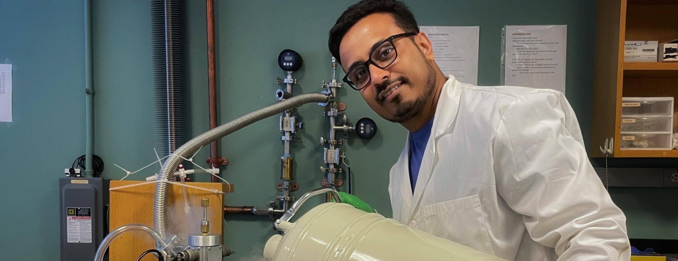 Person in a lab coat and gloves pours material from a large container into a lab apparatus with tubing and gauges.