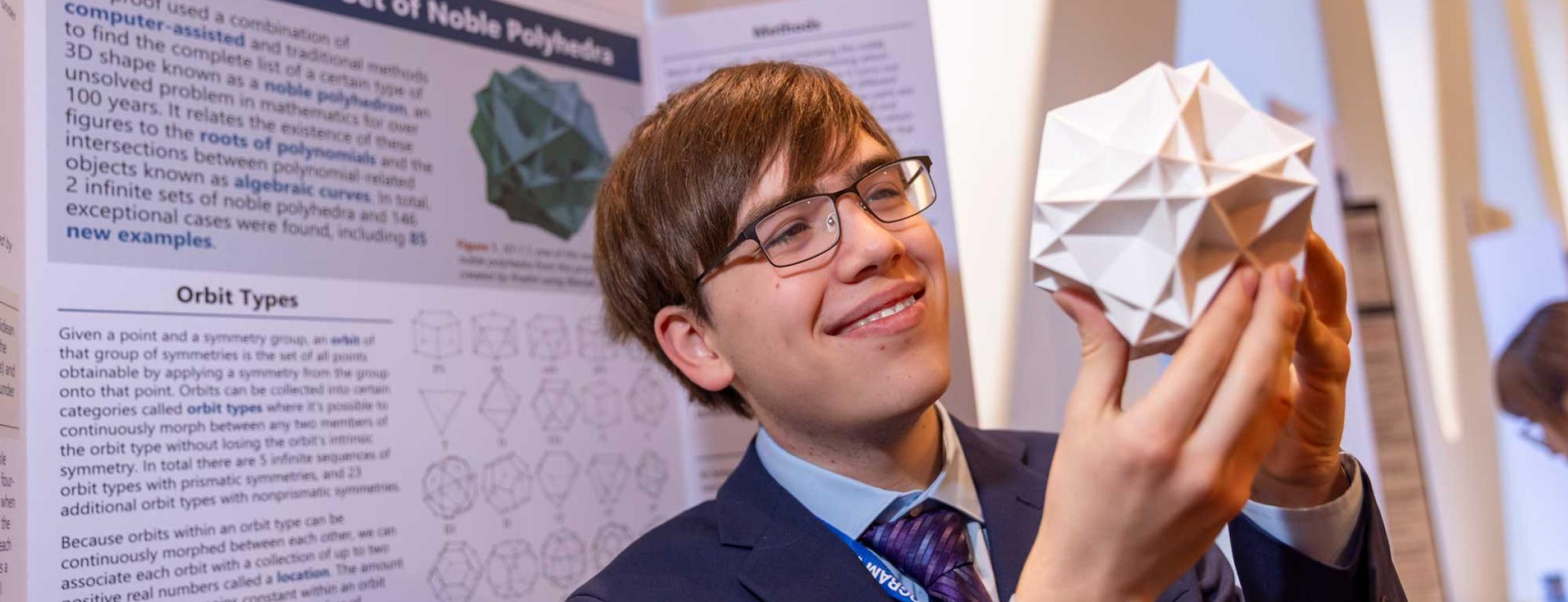 Connor Hill holding model of noble polyhedron in front of his research poster