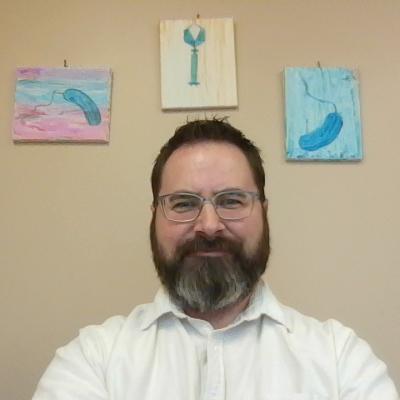 Picture of Dr. Gregory Broussard, white male with beard and white shirt, with paintings of bacteria and phage on the wall behind him.