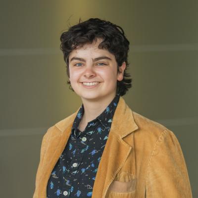 A portrait of Dana Kullgren from the chest up. They are a white person with short, wavy brown hair. They are wearing a black and blue patterned shirt with a tan jacket and smiling at the camera.