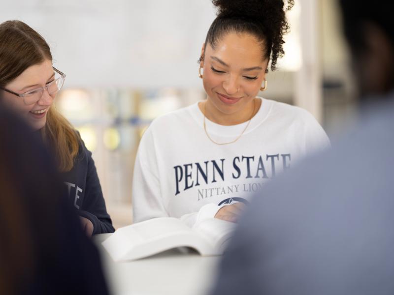 Two coeds, one wearing a Penn State sweatshirt, sit at a table looking at a book and smiling.