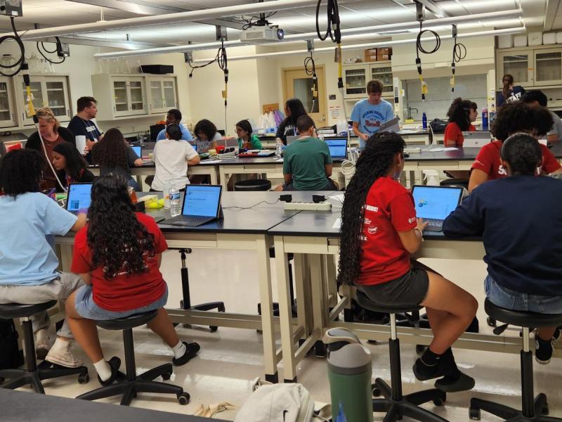 Students working at lab tables with laptops and papers in a bright science classroom.