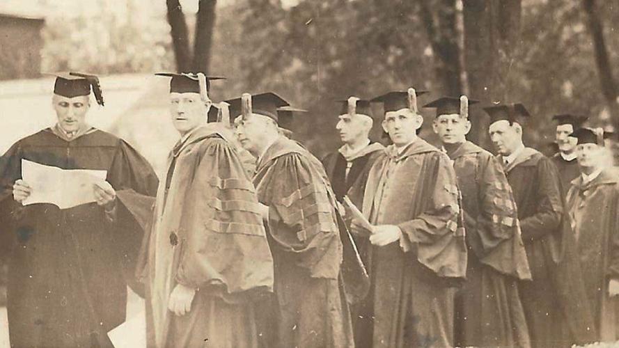 Historical photograph of a group of academics in graduation regalia standing outdoors during a ceremonial procession.