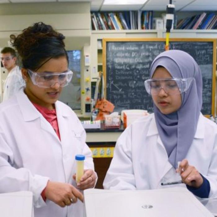 A large group of graduate students in white lab coats working in the laboratory with a chalkboard in the background.