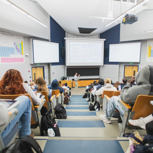 Students sit in a large lecture hall listening to a professor presenting slides at the front of the room.