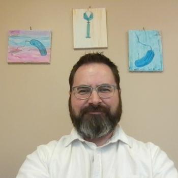 Picture of Dr. Gregory Broussard, white male with beard and white shirt, with paintings of bacteria and phage on the wall behind him.