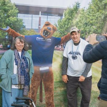 Two attendees of the Penn State Homecoming tailgate posing for a photo with an image of the Penn State Nittany Lion.