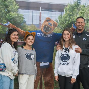 Four people posing in front of an image of the Penn State Nittany Lion at the Homecoming tailgate.