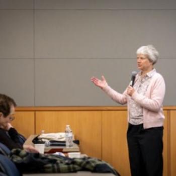 Presenter holding a microphone speaks to an audience in a lecture room, gesturing with one hand near a podium and screen.