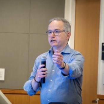 Speaker with microphone gestures forward while presenting beside a computer podium in a classroom setting.