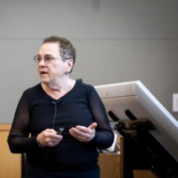 Presenter at podium holds a clicker while speaking during a lecture, standing in front of a screen and equipment.