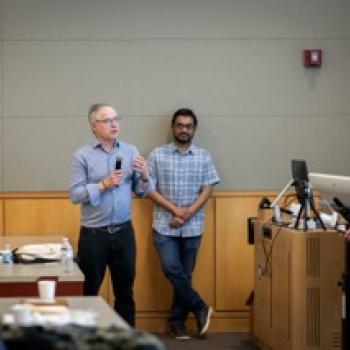 wo speakers stand at front of lecture room; one holds a microphone while another stands beside, audience and podium visible.