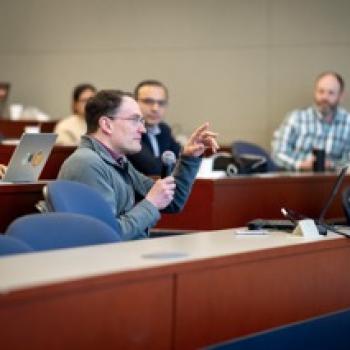 Audience member holds a microphone while speaking from tiered seating, other attendees and laptops visible in classroom.