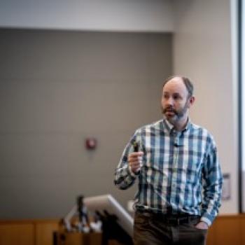 Speaker in checkered shirt holds a remote while presenting beside a projected slide in a lecture room.