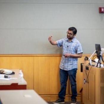 Presenter gestures toward seated attendee while speaking near podium with microphone and equipment on desk.