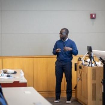 Presenter stands by podium speaking to seated attendee, gesturing while holding a remote in a lecture room.