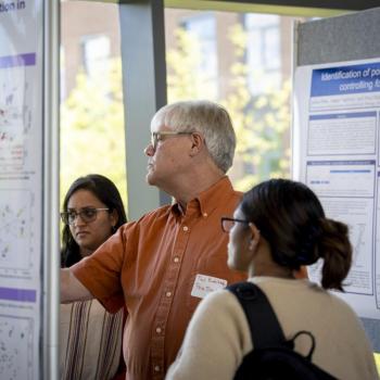 Man points to data on a scientific poster while two attendees stand nearby, examining figures and charts at a research poster session.