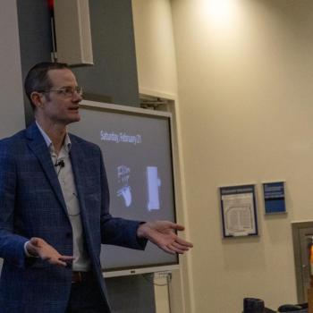 Presenter standing at the front of a lecture hall, gesturing with both hands while speaking beside a projected slide.