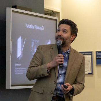 Presenter standing at the front of a lecture hall, gesturing with both hands while speaking beside a projected slide.