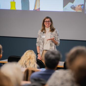 Speaker addressing an audience in a lecture hall, standing in front of a projected slide with images and text.