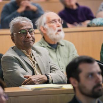 Audience members seated in a lecture hall, watching a presentation, with notebooks and materials on the desks.