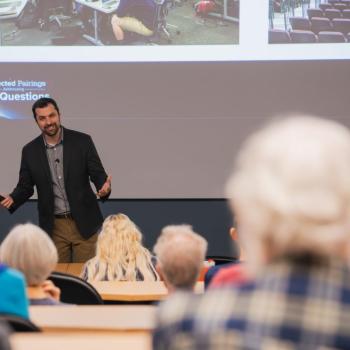 Presenter standing at the front of a lecture hall, gesturing with both hands while speaking beside a projected slide.
