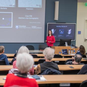 Speaker addressing an audience in a lecture hall, standing in front of a projected slide with images and text.