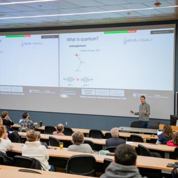 Speaker addressing an audience in a lecture hall, standing in front of a projected slide with images and text.