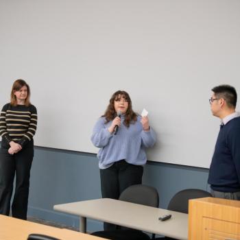 Three people standing at the front of a classroom, with one holding a microphone and speaking while the others listen.