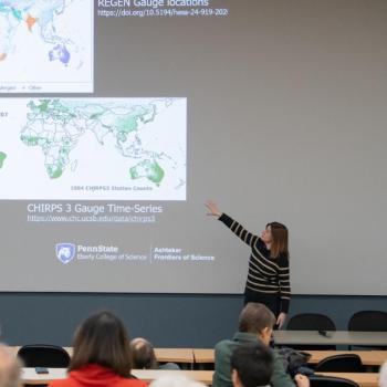 Speaker addressing an audience in a lecture hall, standing in front of a projected slide with images and text.