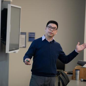 Presenter standing at the front of a lecture hall, gesturing with both hands while speaking beside a projected slide.