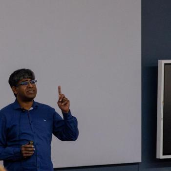 Presenter standing in front of a large projected screen, holding a small device and raising one hand while addressing an audience.