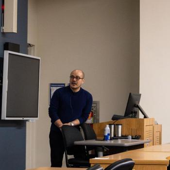 Speaker addressing an audience in a lecture hall, standing in front of a projected slide with images and text.