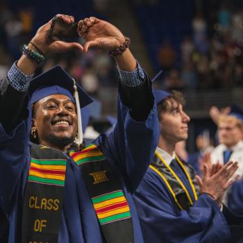A new graduate makes a heart symbol with his hands directed at his family in the audience.