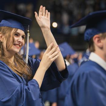 A standing coed students waves to her family in audience towards the end of the ceremony.