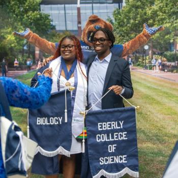 A future graduate and family pose with commencement banners in front of the Nittany Lion Mascot backdrop.