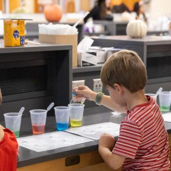 Child in a striped shirt uses a dropper to add liquid to clear cups filled with colored solutions on a lab counter. Additional cups, droppers, and pumpkins are visible in the background.