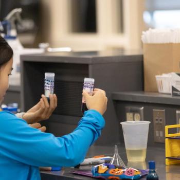 Two individuals at a lab counter hold test tubes with layered colored substances. Nearby are a clear plastic cup, funnels, and Halloween-themed decorations.