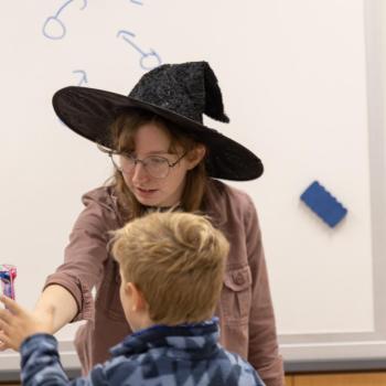 Person wearing a black witch hat hands a colorful item to a child in front of a whiteboard with blue marker drawings of circles and arrows.