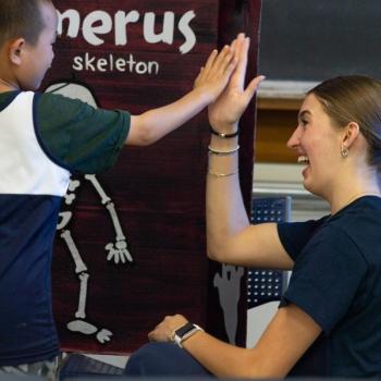 Child and adult give each other a high-five in front of a dark red display featuring a skeleton graphic and the words “in the humerus skeleton.” The background includes a chalkboard and classroom furniture.