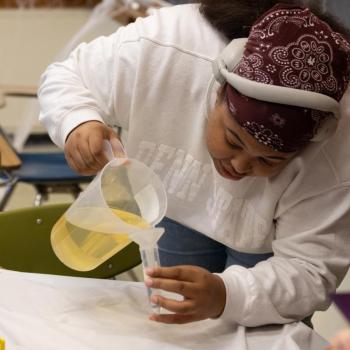 Person wearing a white sweatshirt and patterned head covering pours yellow liquid from a large plastic pitcher into a smaller graduated cylinder on a table covered with a white cloth. Small colorful objects and straws are scattered on the table, and a chalkboard with faint writing and cobweb decorations is in the background.