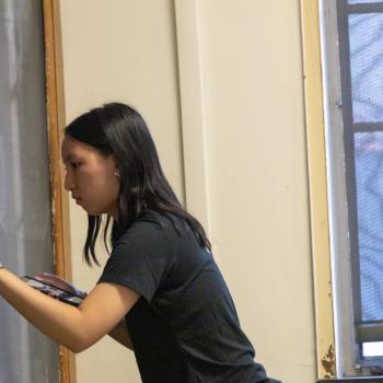 Person in a black shirt draws a ghost figure on a chalkboard using white chalk. A spiderweb design is already drawn on the board, and yellow caution tape decorates the edges. The person holds a small container of chalk pieces in one hand while sketching with the other. A window with a mesh screen is visible in the background.