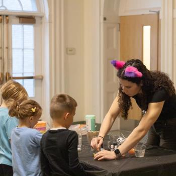 Individual with colorful cat ear headband leans over a black-covered table, assisting children with a hands-on activity involving clear cups and small containers. The setting appears to be a hallway or classroom with wooden doors and arched windows in the background.