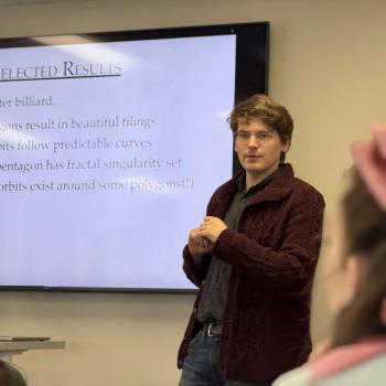 Presenter standing near a screen displaying a slide titled “Selected Results” with bullet points about polygons and orbits.