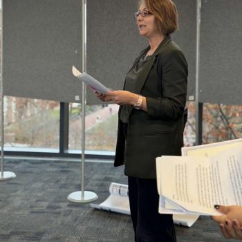 Person standing indoors holding papers, with large windows and gray display boards in the background.