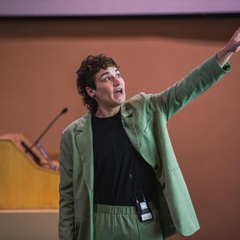 Presenter stands at a podium area, arm raised and pointing toward a projection screen in a lecture hall.