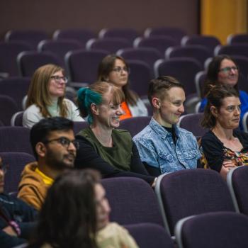 Audience members sit in tiered seats, facing forward and listening during a talk in a lecture hall.