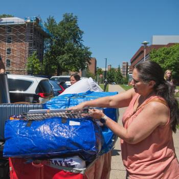 Students and family loading blue bags, a suitcase and a trash bin into a pickup truck during a sunny college move-in, with a brick dorm wrapped in scaffolding visible behind them.