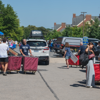 Students and families unloading cars during college move-in day, pushing red rolling bins and carrying boxes and bags along a sunny street lined with parked cars, trees, and campus buildings.