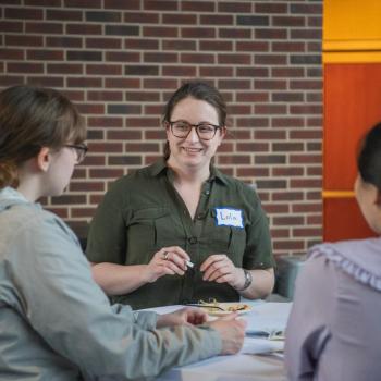 Three people sit at a table talking during a networking event, with papers and snacks in front of a brick wall.